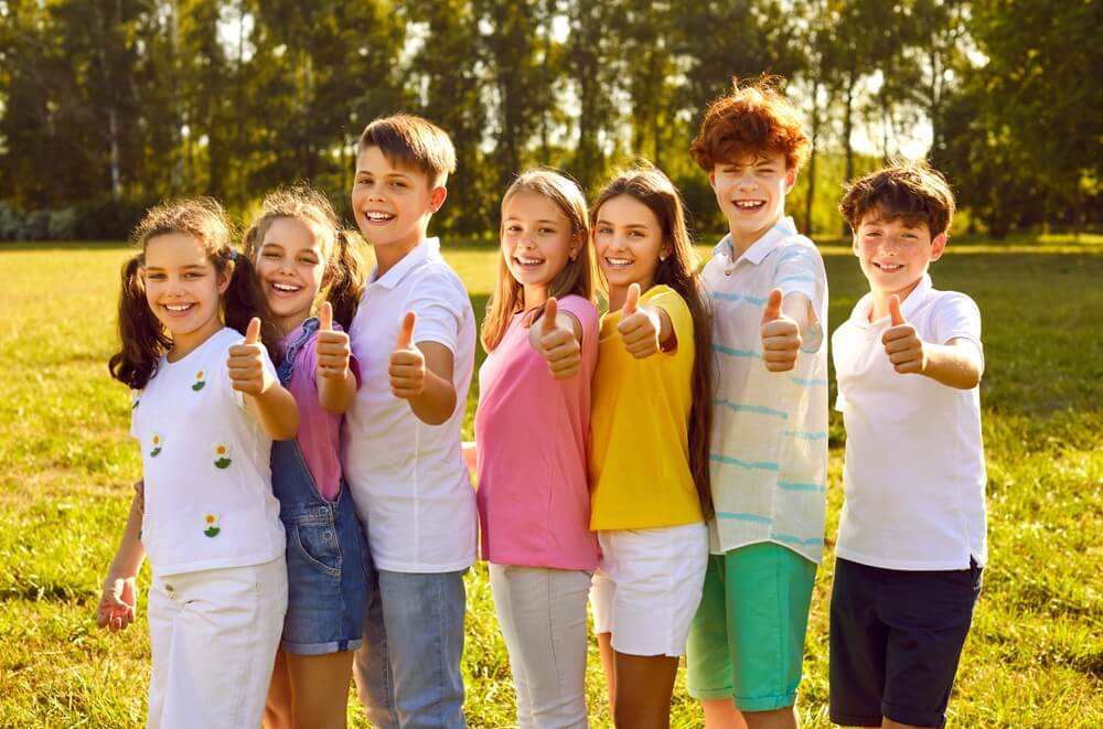 Smaidoši bērni dažādās vecuma grupās stāv zaļā parkā saulainā dienā, skatās kamerā un rāda īkšķus uz augšu, simbolizējot draudzību, pozitīvu pieredzi un aktīvu dalību bērnu vasaras nometnēs 2026
