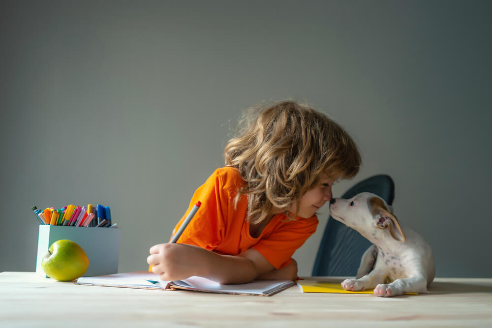 Bērns sēž pie galda un raksta burtnīcā, kamēr mazs suns pieskaras viņa sejai, radot rotaļīgu un emocionāli pozitīvu vidi, kas simbolizē dabisku valodu apguvi caur ikdienas situācijām un komunikāciju bērnu nometnēs